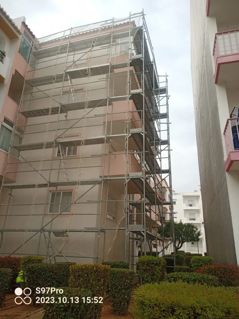 Edifício residencial de cor salmão totalmente coberto por andaimes e uma rede de proteção, indicando trabalhos de reabilitação de fachadas.