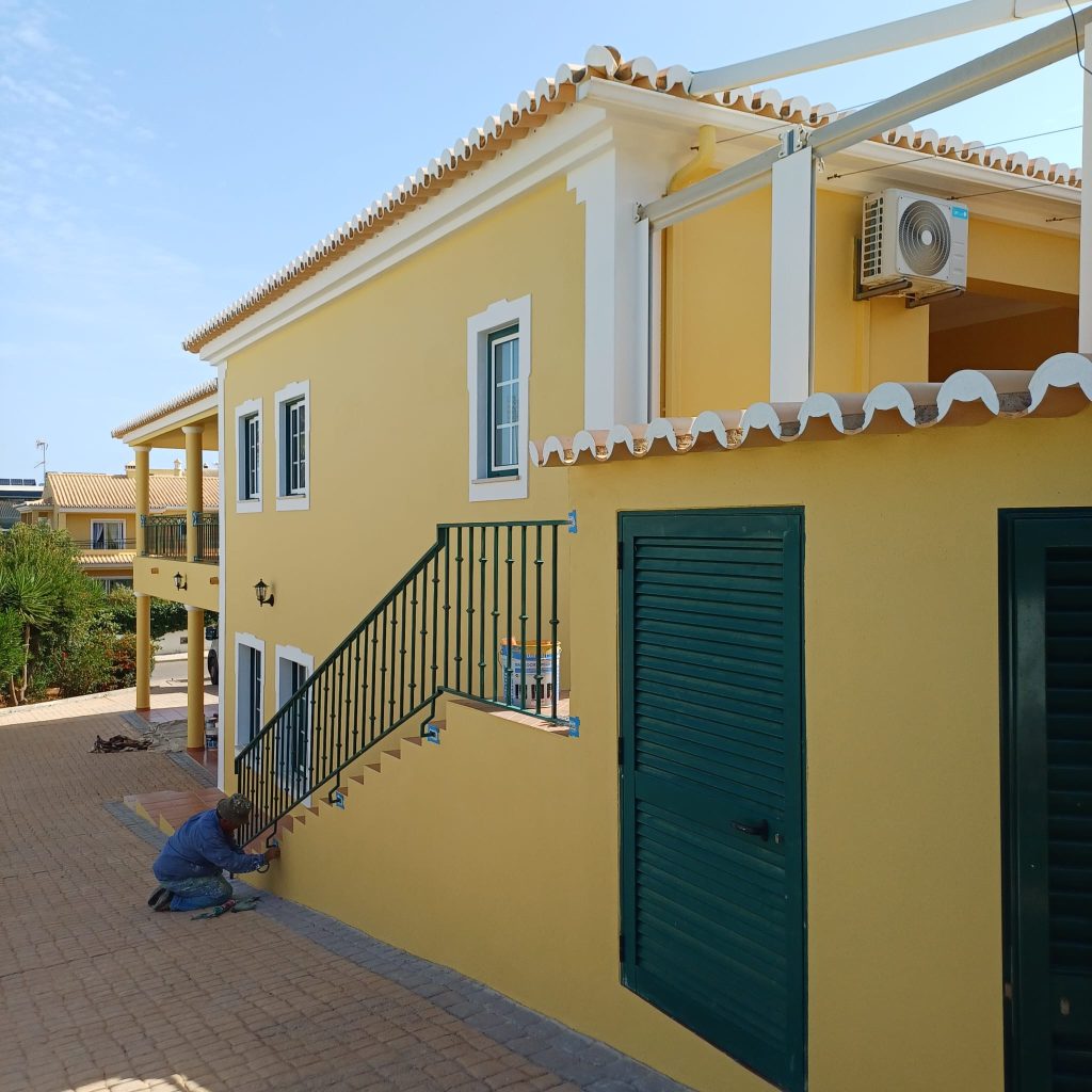 Pintor profissional a realizar acabamentos de pintura na base de um muro de escadas exteriores de uma moradia amarela. A casa apresenta detalhes em branco, janelas de caixilharia verde e telhado tradicional português.