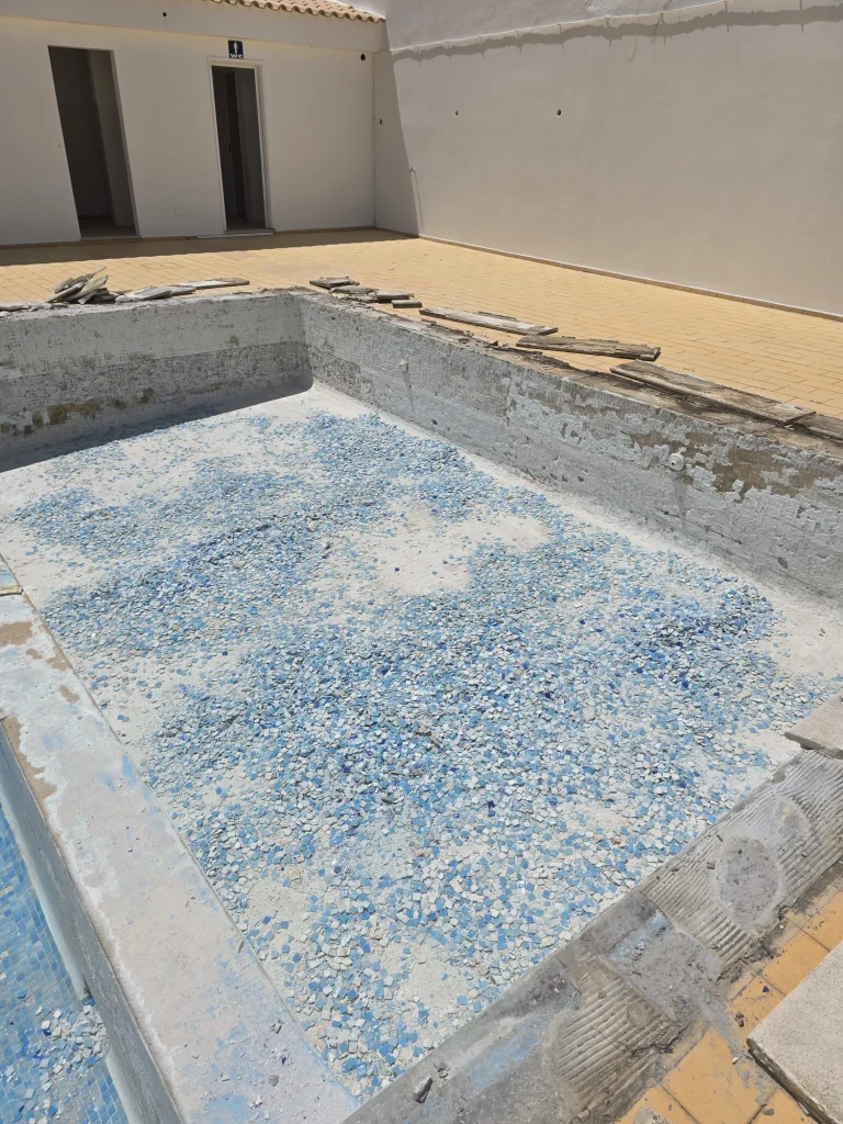 Piscina vazia em obras de reconstrução, com o fundo coberto de fragmentos de pastilhas azuis e brancas removidas, e pavimento circundante amarelo.