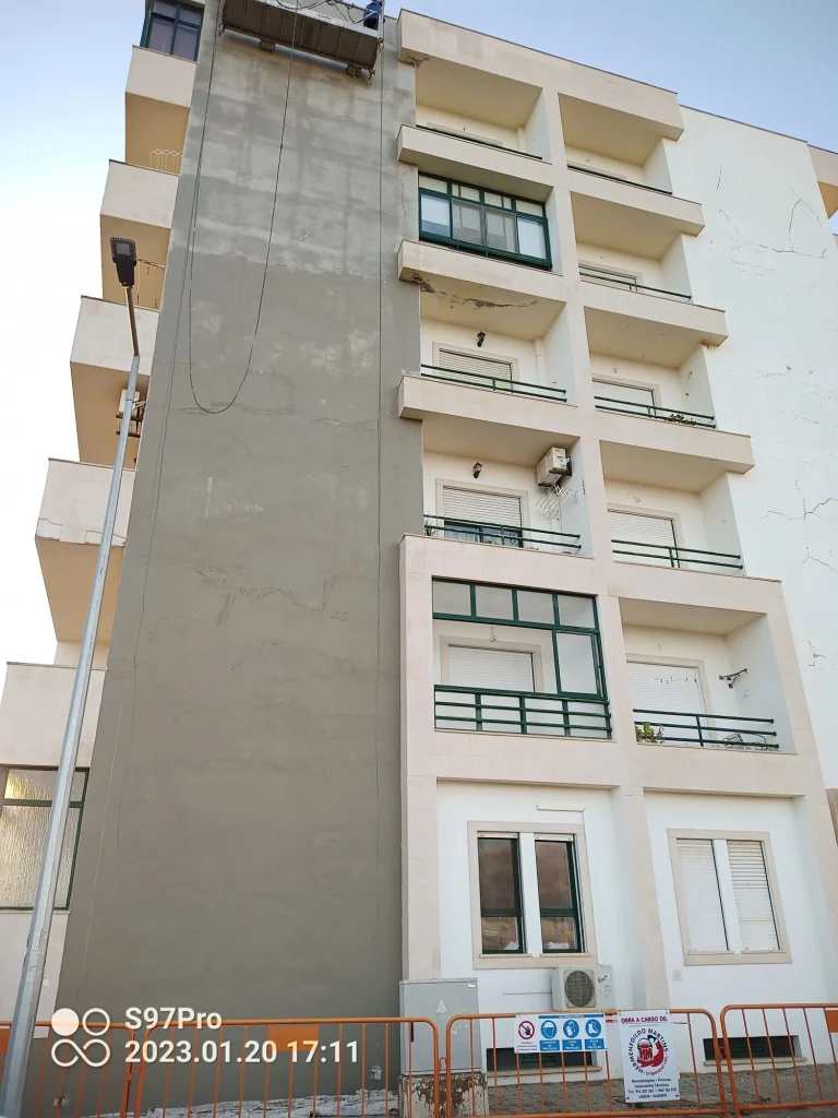 Fachada de edifício residencial em reabilitação, mostrando uma grande área com aplicação de argamassa ou betonilha cinzenta, com rachas visíveis na secção branca.
