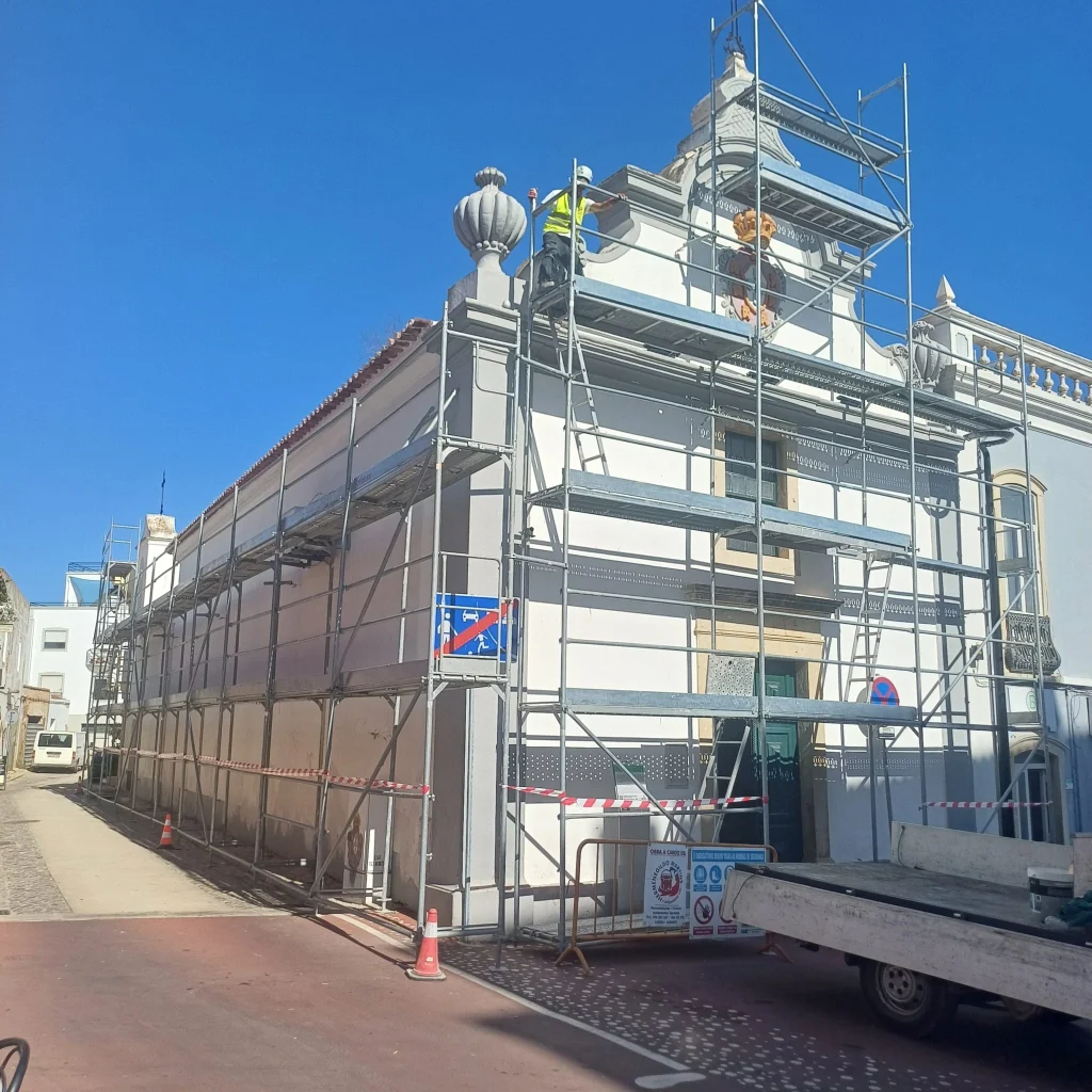 Andaime montado na fachada de um edifício antigo ou igreja, com elementos decorativos no topo, indicando obras de restauro e preservação do património.