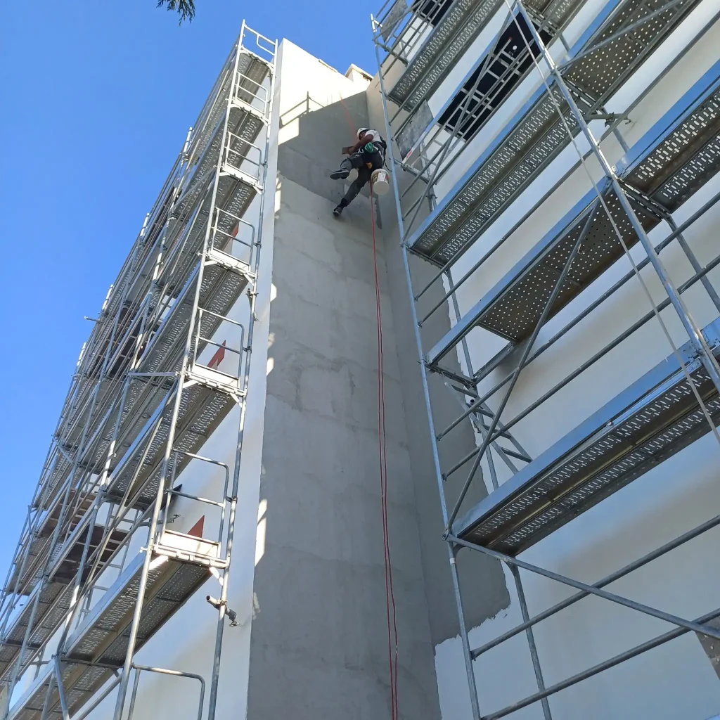 Trabalhador especializado em trabalhos verticais realiza a reabilitação de uma fachada de prédio, suspenso por cordas de segurança entre duas estruturas de andaimes metálicos sob um céu azul.
